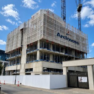 Under-construction multi-story building with scaffolding and a crane overhead against a blue sky with clouds, safety barriers along the street.