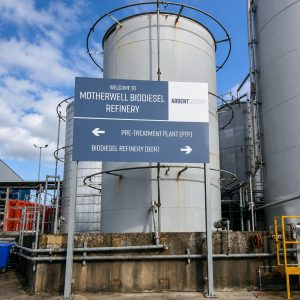 Sign at a biodiesel refinery directing to Pre-Treatment Plant left and Biodiesel Refinery right; large blue sign mounted on metal posts among gray industrial tanks with piping around. (Informative)