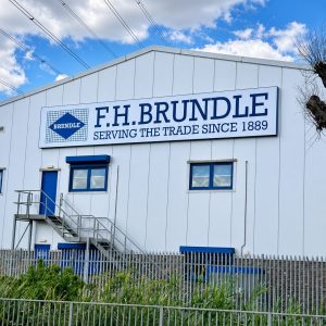 Industrial building exterior with a large F.H. Brundle sign reading 'Serving the Trade Since 1889', blue doors, metal staircase, and a tall metal fence in the foreground against a blue sky with clouds.