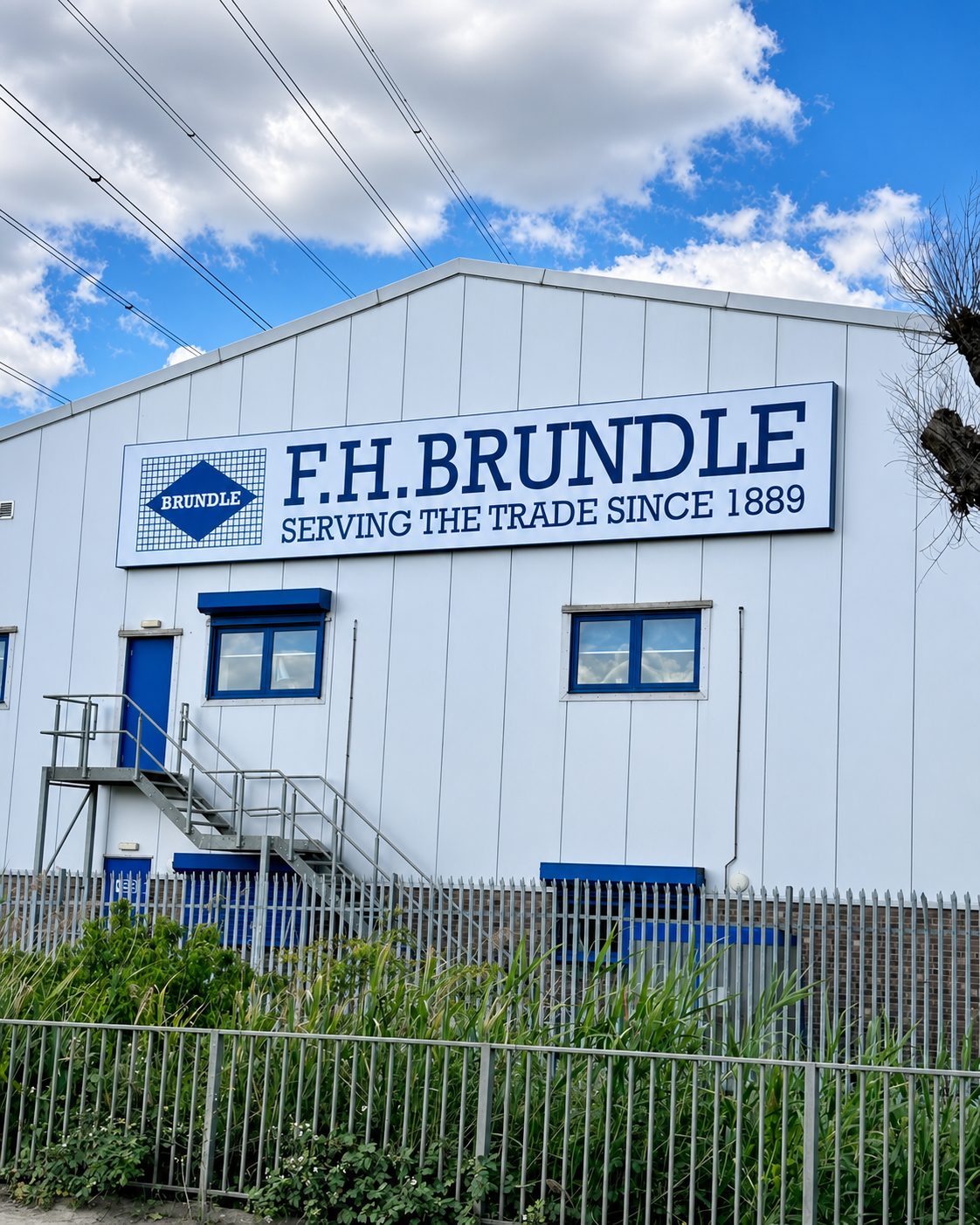 Industrial building exterior with a large F.H. Brundle sign reading 'Serving the Trade Since 1889', blue doors, metal staircase, and a tall metal fence in the foreground against a blue sky with clouds.