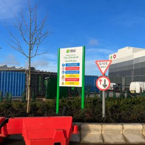 Tall green-framed sign listing yard rules outside an industrial site, with a give-way sign and red plastic barriers in the foreground.