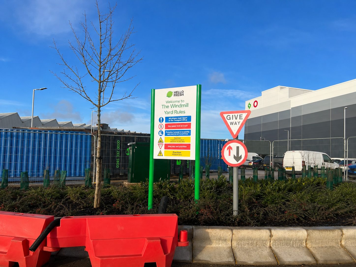 Tall green-framed sign listing yard rules outside an industrial site, with a give-way sign and red plastic barriers in the foreground.