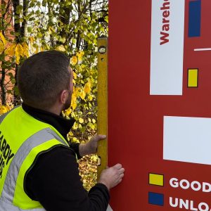 Worker in a bright safety vest attaches a large red signboard to a wooden post outdoors, autumn leaves in the background.