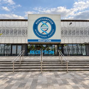 Front facade of Northampton International Academy reception with a large circular logo, blue 'RECEPTION' sign, and glass doors.