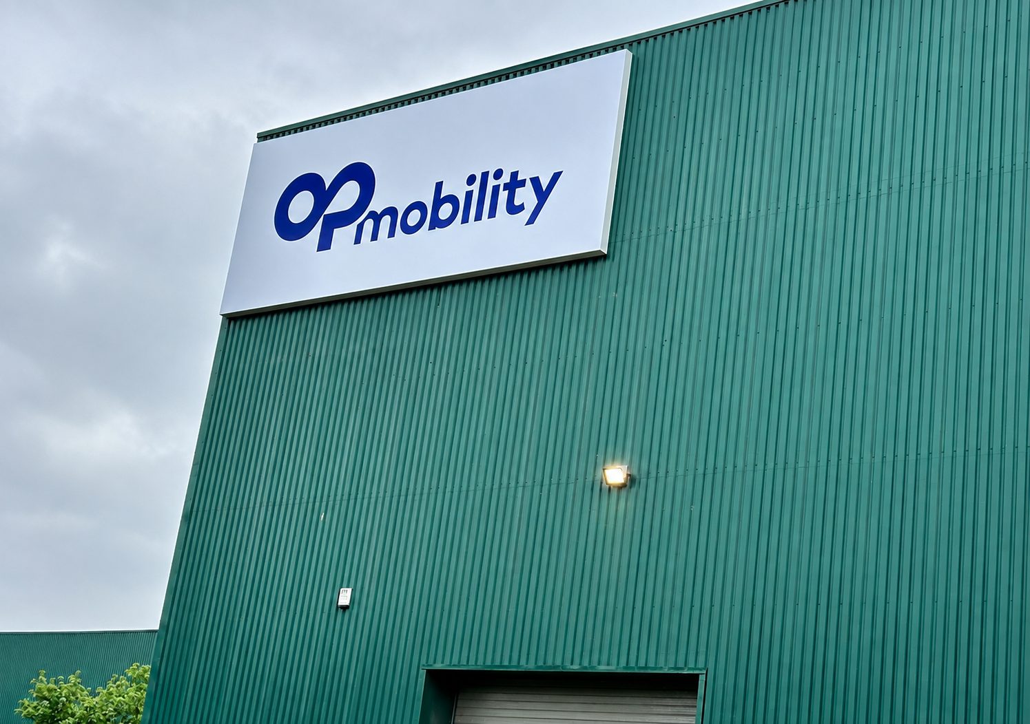 Green industrial building with a large white sign reading 'Op mobility' in blue, under a cloudy sky.