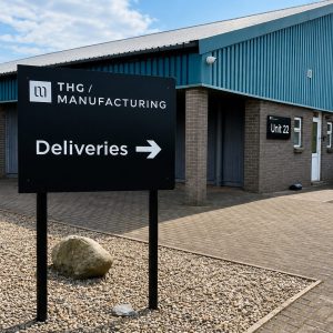 Exterior of THG Manufacturing building with a large black 'Deliveries' sign and arrow, Unit 22, blue metal roof and brick walls.