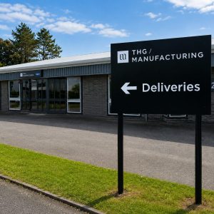 Exterior view of THG Manufacturing with a large black sign reading Deliveries and a left-pointing arrow.