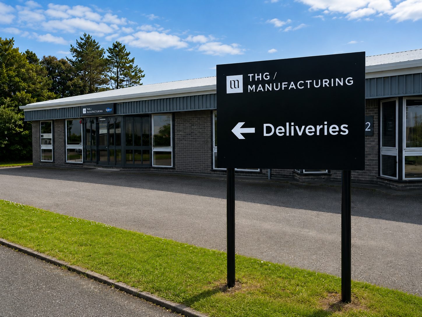 Exterior view of THG Manufacturing with a large black sign reading Deliveries and a left-pointing arrow.