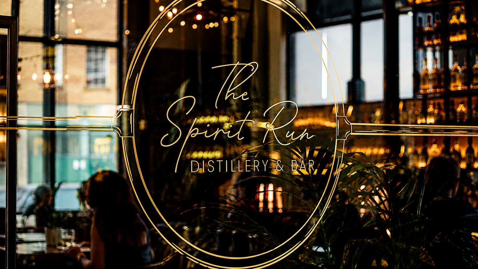 Window sign reading 'The Spirit Run Distillery & Bar' in a circular gold emblem with warm interior lights behind glass.