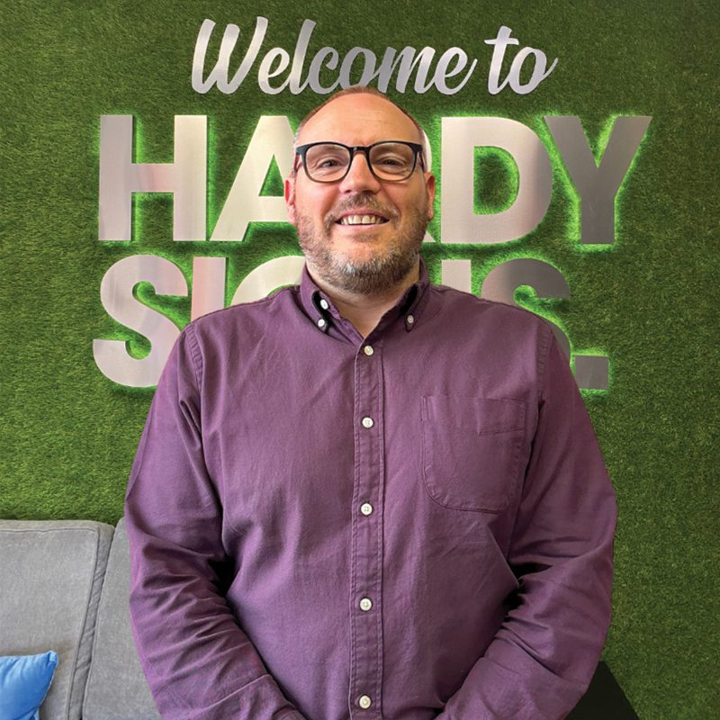 Smiling man with glasses and a purple button-up shirt stands in front of a green wall with large white text reading “Welcome to …” behind him.