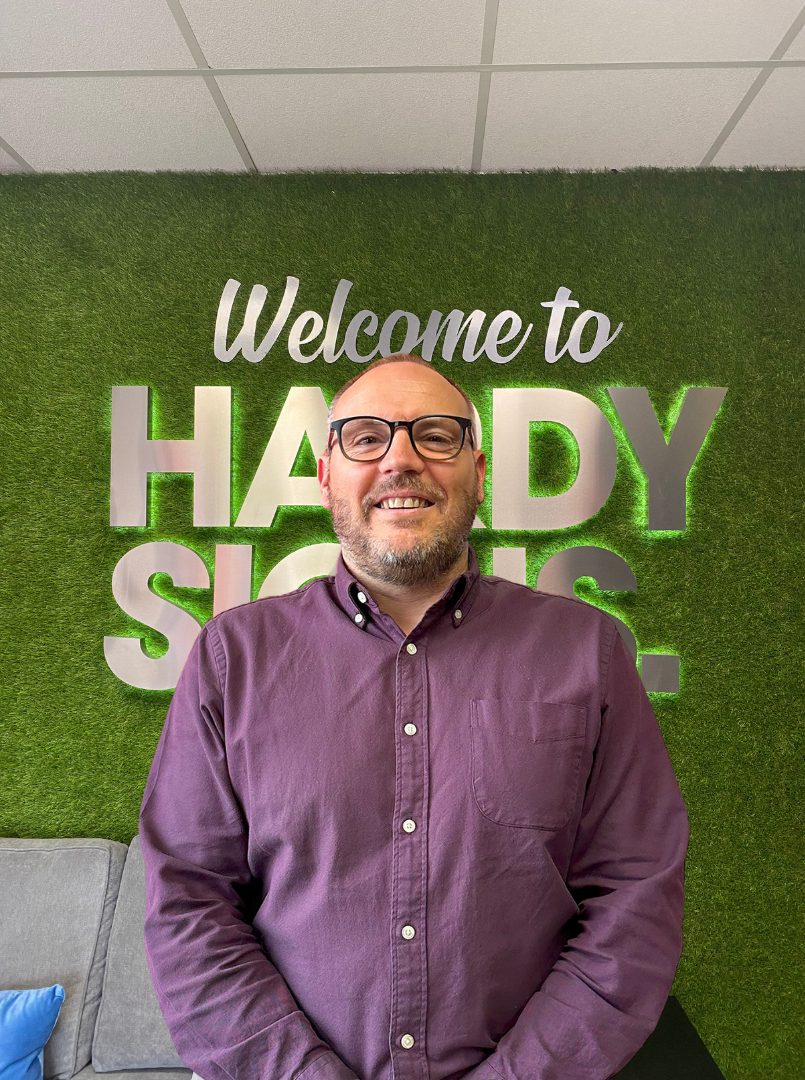 Smiling man in a purple shirt and glasses stands in front of a green faux-grass wall with a 'Welcome to HARDY SHOPS' sign in the background.