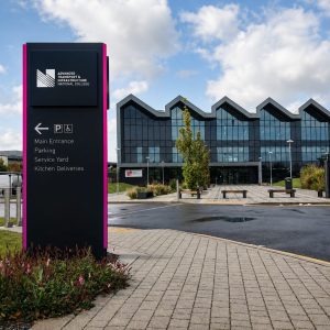 Tall black directional sign with magenta edges for Advanced Transport & Infrastructure National College; lists Main Entrance, Parking, Service Yard, Kitchen Deliveries, with a modern glass-front campus building in the background.