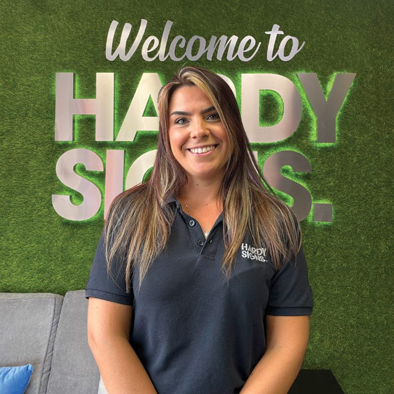 Smiling woman in a dark polo shirt stands in front of a green wall with large 'HARDY SIGNS' lettering and 'Welcome to' at the top.