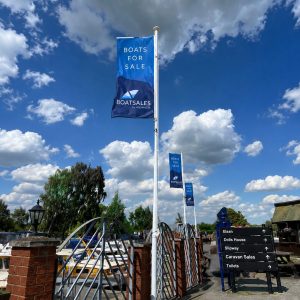 Blue sky with scattered clouds at a marina; tall flag banners read 'BOATS FOR SALE' near boats and a signpost at right.