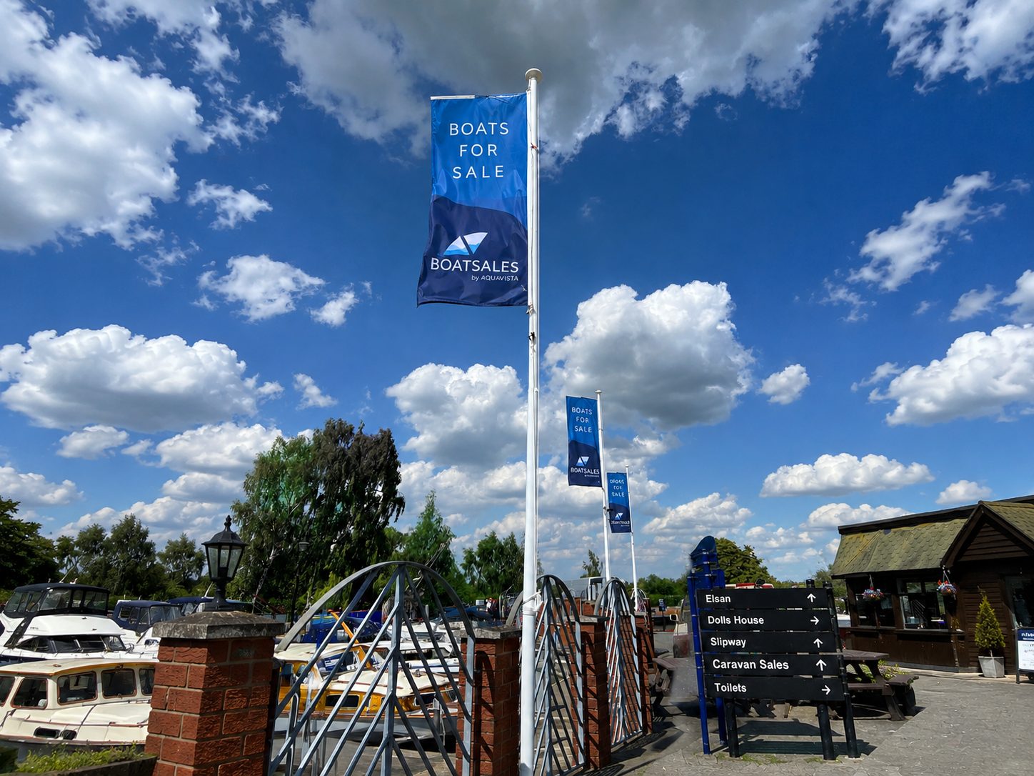 Blue sky with scattered clouds at a marina; tall flag banners read 'BOATS FOR SALE' near boats and a signpost at right.
