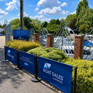 Boats docked along a marina behind blue Boat Sales banners and decorative brick pillars on a sunny day.",
