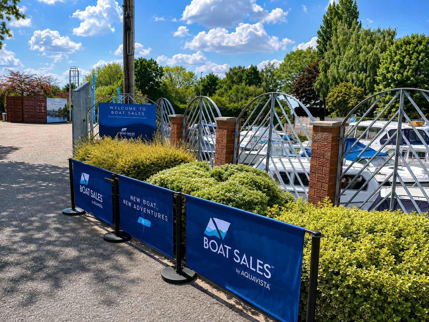 Boats docked along a marina behind blue Boat Sales banners and decorative brick pillars on a sunny day.",