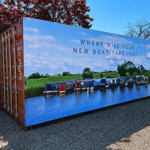 Billboard on a shipping container showing a row of canal boats on a river with the caption 'Where will your new boat take you?'.