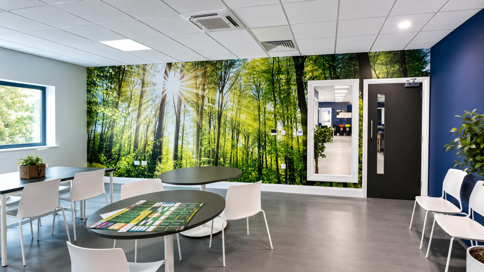 Bright lounge area with a forest mural wall, round black tables and white chairs, and a blue accent wall with plants nearby.