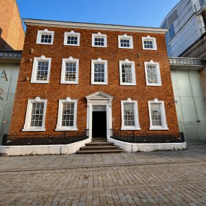 Brick three-story building with white-framed windows and a central doorway reached by steps, framed by a black railing on a cobblestone street.