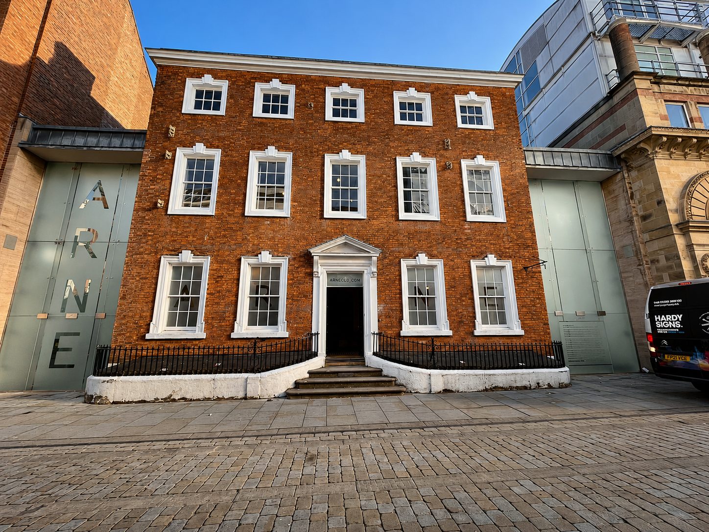 Brick three-story building with white-framed windows and a central doorway reached by steps, framed by a black railing on a cobblestone street.
