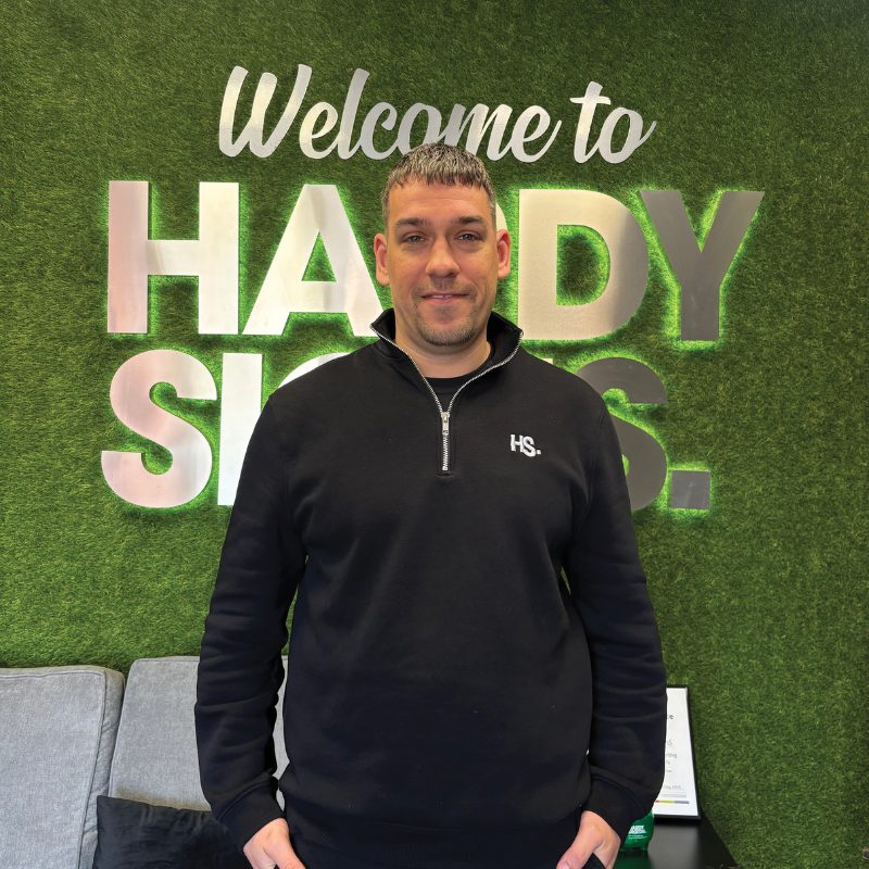 Man in a black pullover poses in front of a green turf wall with large metallic letters forming a sign behind him (text partially reads 'Welcome to').