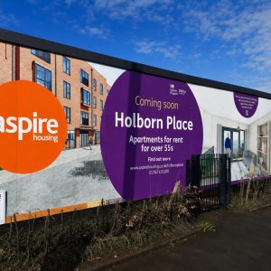 Advertisement boards on a construction fence: Aspire Housing logo in an orange circle and a purple panel announcing Holborn Place apartments for rent for over 55s, with a street view in the background.