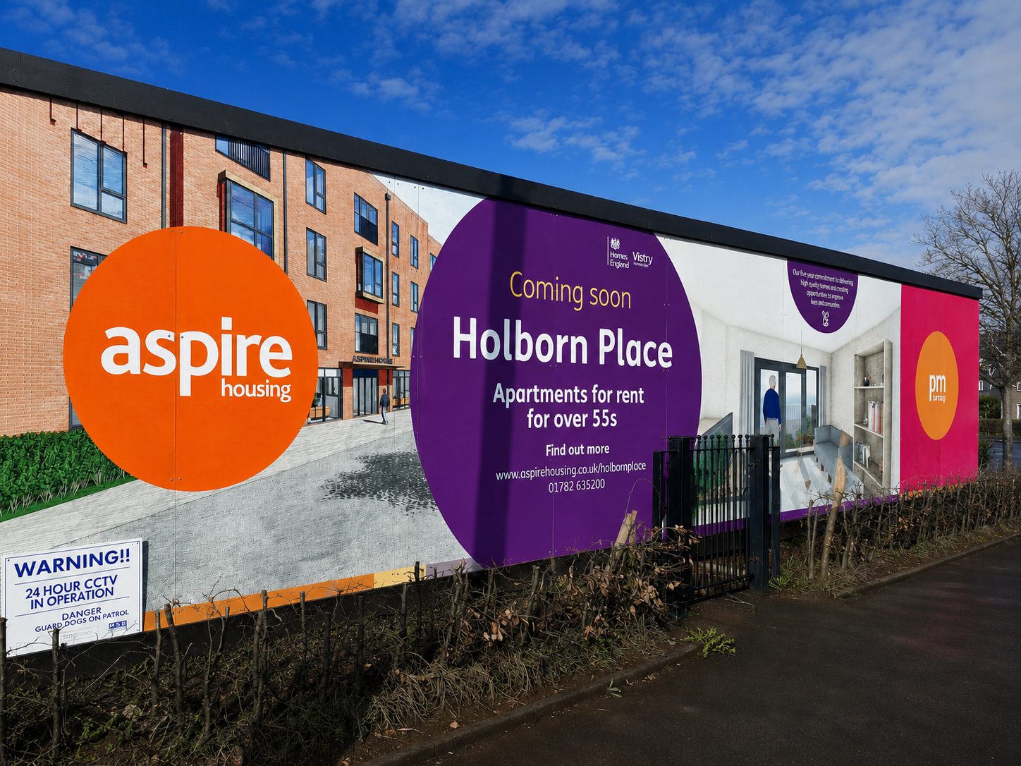 Advertisement boards on a construction fence: Aspire Housing logo in an orange circle and a purple panel announcing Holborn Place apartments for rent for over 55s, with a street view in the background.