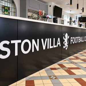 Reception desk at Aston Villa Football Club, with bold white 'ASTON VILLA FOOTBALL CLUB' lettering on a black panel and a patterned tiled floor behind. The bar area and pendant lights are visible above the counter.