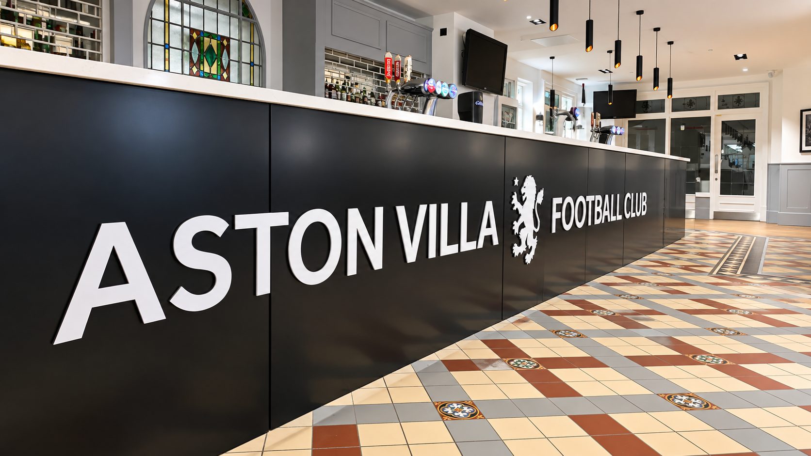 Reception desk at Aston Villa Football Club, with bold white 'ASTON VILLA FOOTBALL CLUB' lettering on a black panel and a patterned tiled floor behind. The bar area and pendant lights are visible above the counter.