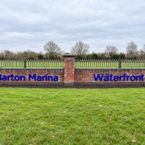 Brick sign reading 'Barton Marina' and 'The Waterfront' in blue letters along a low brick wall with string lights, green lawn in front and trees in the background.