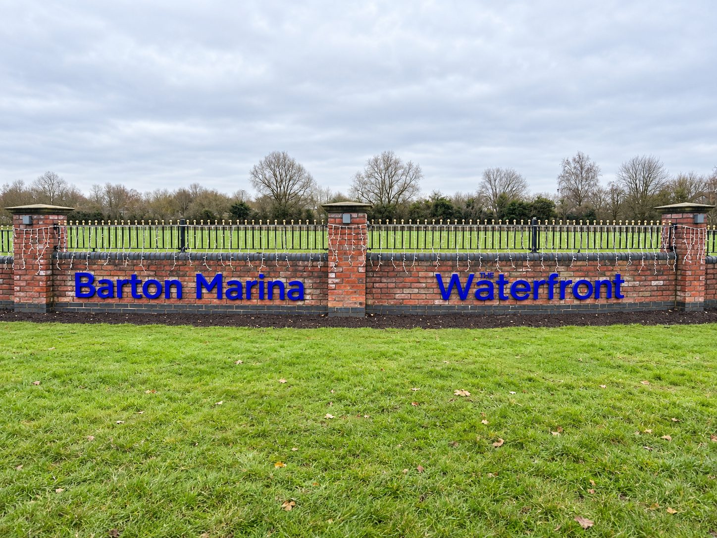 Brick sign reading 'Barton Marina' and 'The Waterfront' in blue letters along a low brick wall with string lights, green lawn in front and trees in the background.
