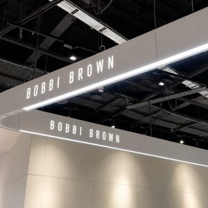 Overhead curved sign reading BOBBI BROWN in bright letters above a neutral retail booth.