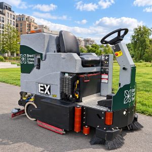 Ride-on street sweeper parked on a paved park path with modern brick apartment buildings in the background.