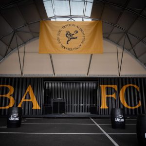 Dimly lit indoor arena with a large yellow Burton Albion banner hanging from the ceiling, orange letters on the back wall, and CARLING-branded punching bags in the foreground.