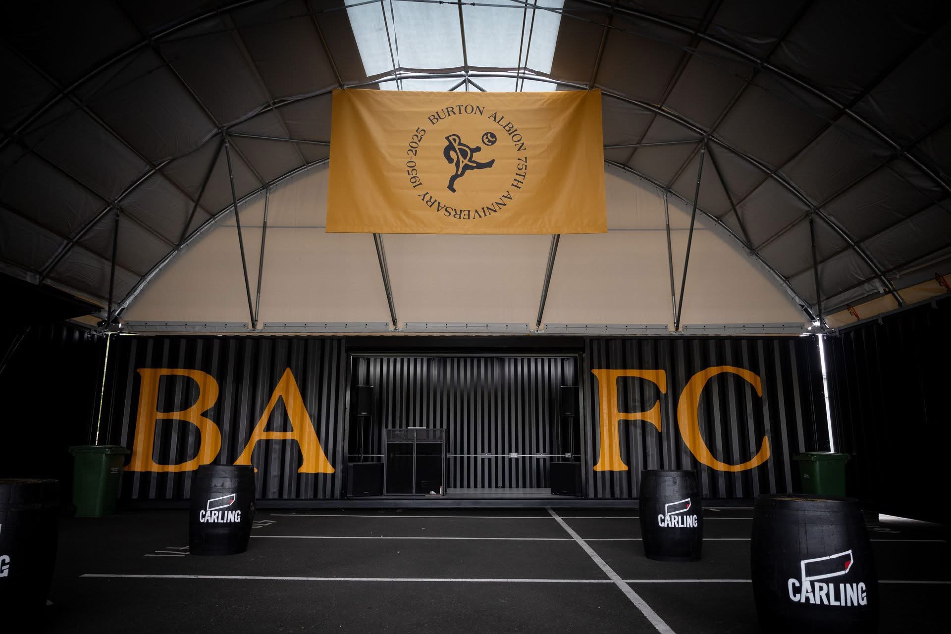 Indoor event space with a large yellow Burton Albion banner hanging from a curved ceiling and orange BARTEC letters on black walls behind several Carling-barrel barriers.