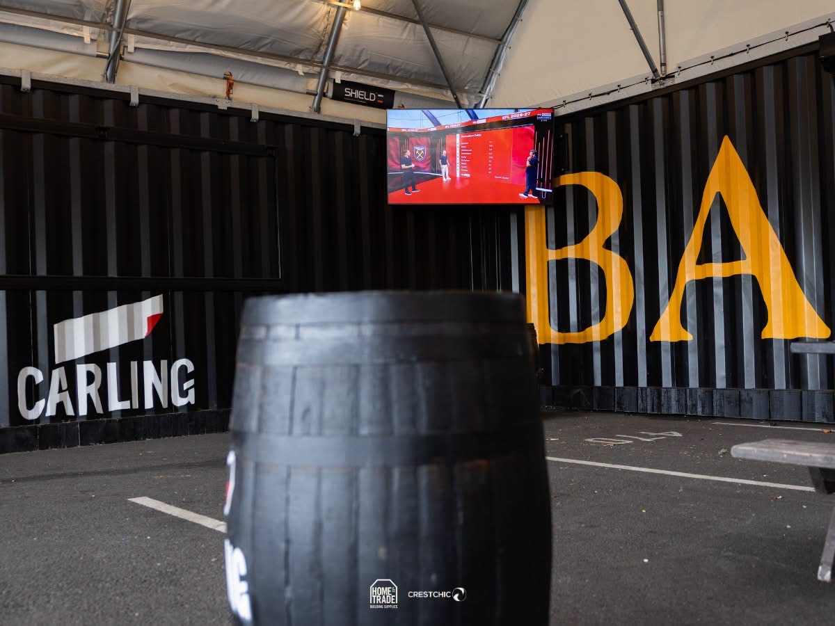 Barrel in the foreground at an indoor event space, with a dark container wall showing a large yellow 'BA' and a Carling sign; a TV screen hangs overhead.