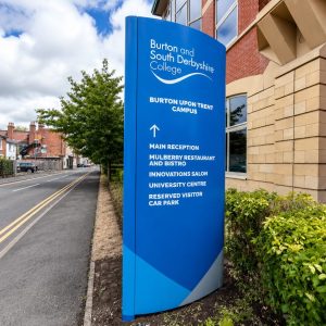 Blue campus sign for Burton and South Derbyshire College with a white wave logo and directions to main reception and facilities.