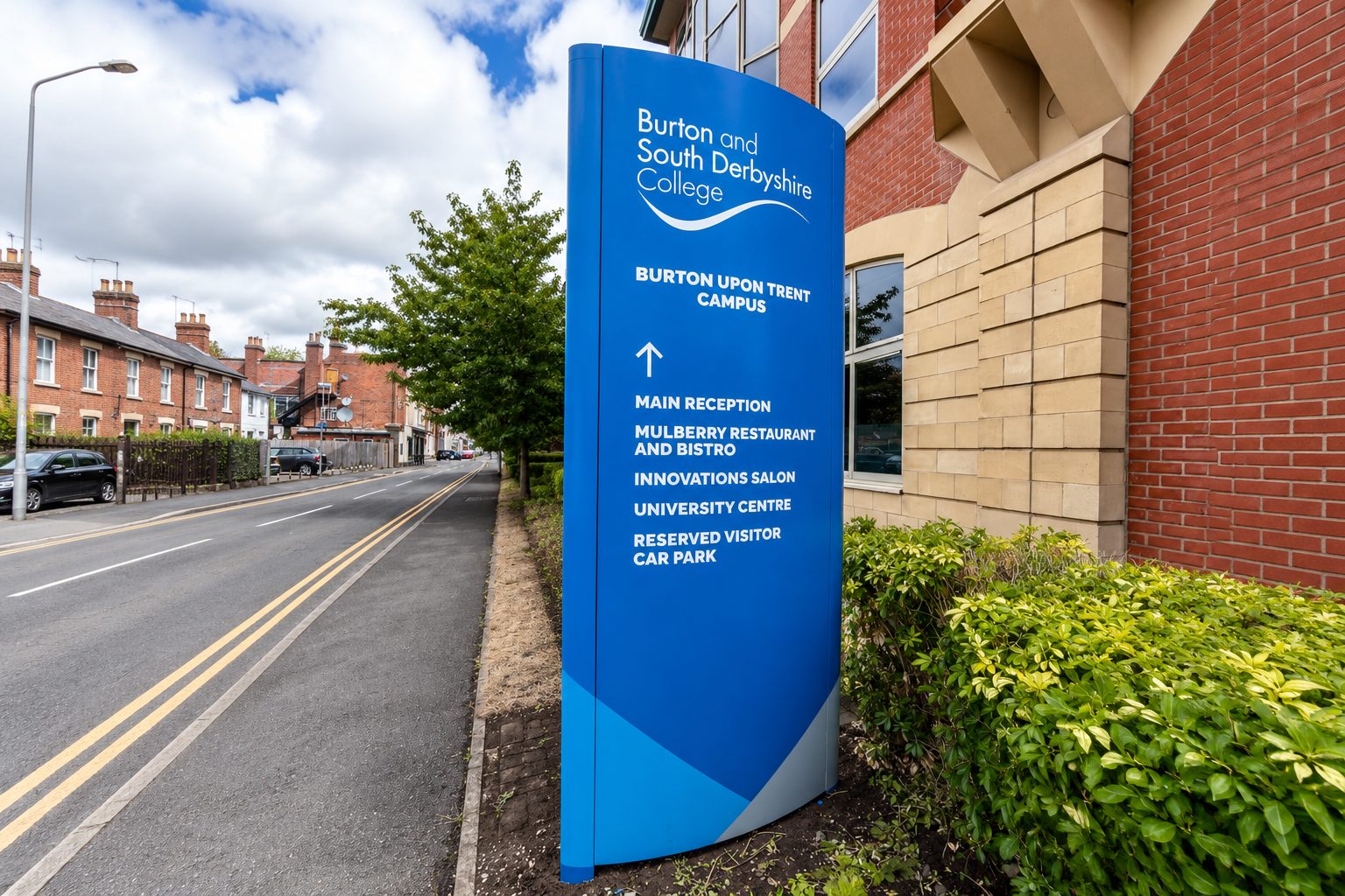 Blue campus sign for Burton and South Derbyshire College with a white wave logo and directions to main reception and facilities.