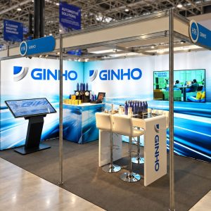 GINHO trade show booth with blue branding, large backlit panels reading GINHO, touchscreen kiosk, and white bar-height seating area at the booth.
