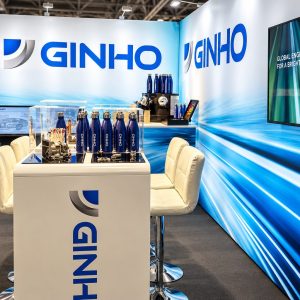 Trade-show booth for GINHO with a white counter, blue bottled products, seating, and a large blue GINHO logo on the backdrop.