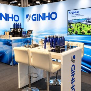 Ginho trade show booth with blue gradient walls, large GINHO logos, and white high table with chairs displaying bottles and sample items.