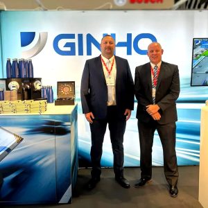 Two men in suits pose at a trade show booth with a blue branded backdrop reading 'GINHO' and product displays.