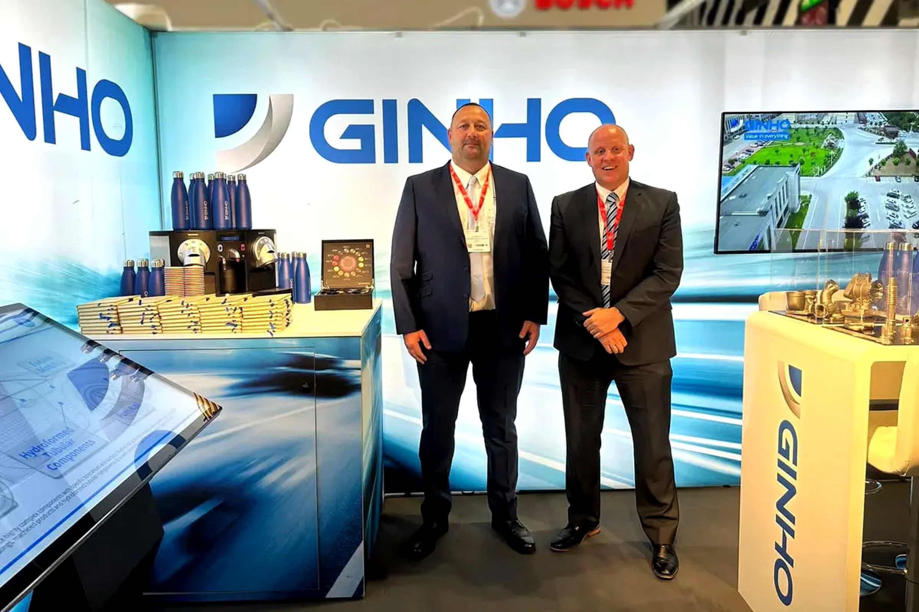 Two men in suits pose at a trade show booth with a blue branded backdrop reading 'GINHO' and product displays.