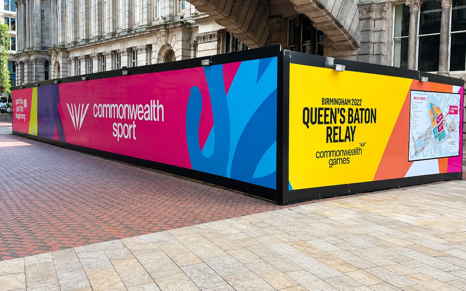 Colorful barrier with Birmingham 2022 Queen's Baton Relay and Commonwealth Games branding, set against a historic building exterior.