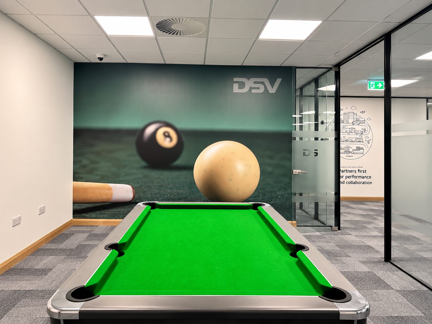 Pool table in a modern office recreation area with a large billiard ball and cue against a DSV-branded wall.