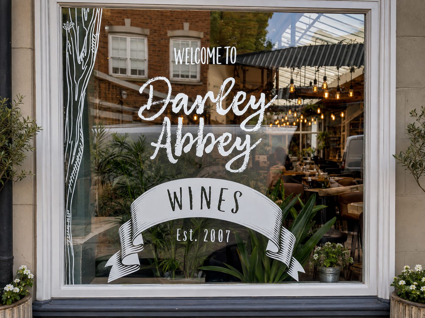 Storefront window sign reading 'Welcome to Danley Abbey Wines Est. 2007' with a decorative ribbon, plants, and a cozy indoor dining area visible through glass.