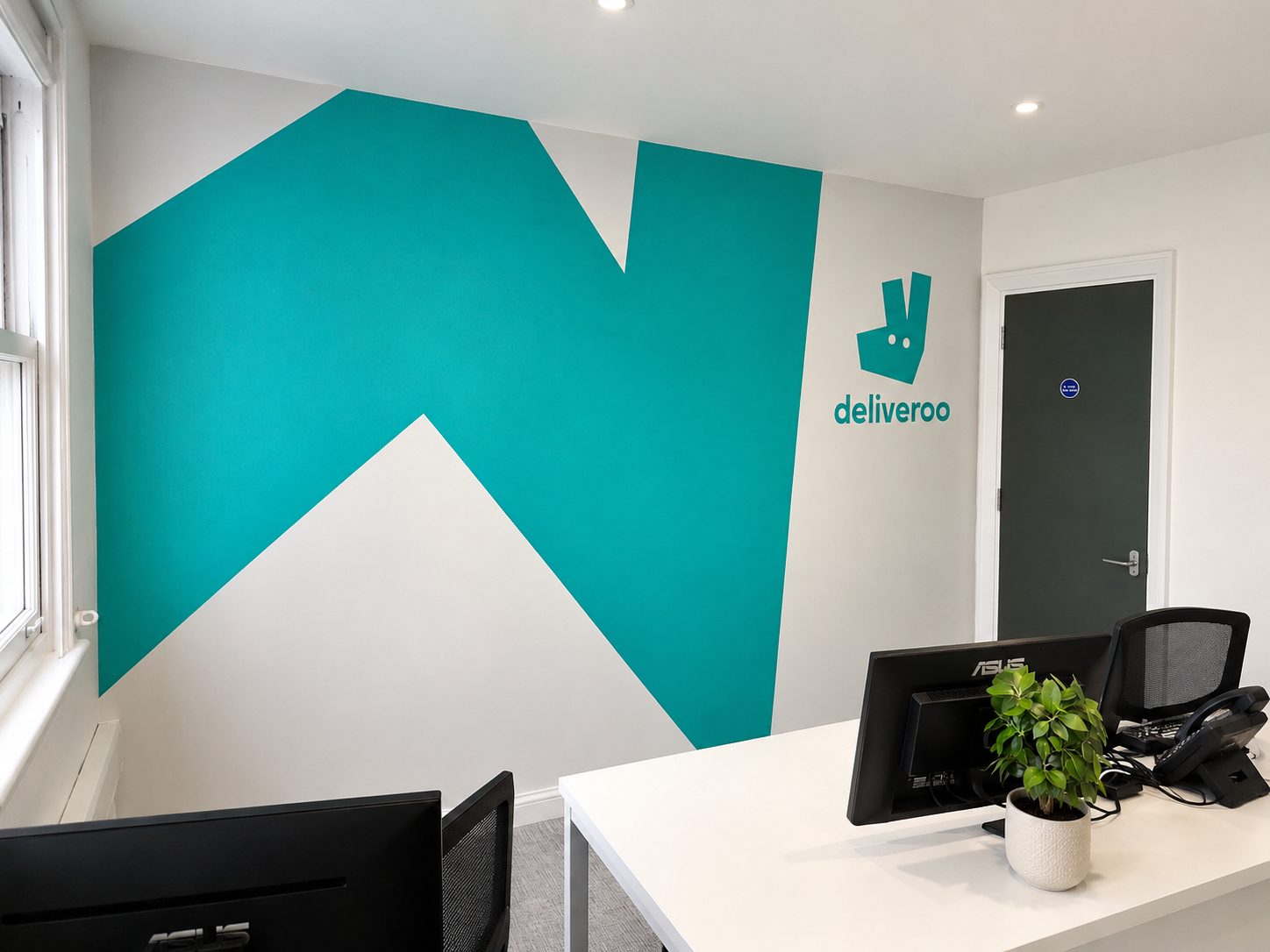 Office with teal geometric wall mural and Deliveroo logo, two desks and computer monitors in foreground