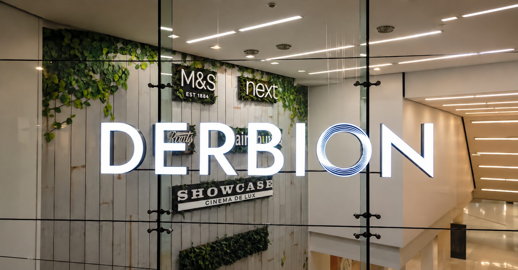 Illuminated storefront sign reading 'DERBION' on a glass wall with greenery and a white-washed backdrop in a mall corridor