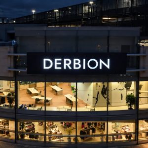 Night exterior of a curved glass shopping center with a bright DERBION sign; interior desks, tables, and clothing racks visible through the windows.